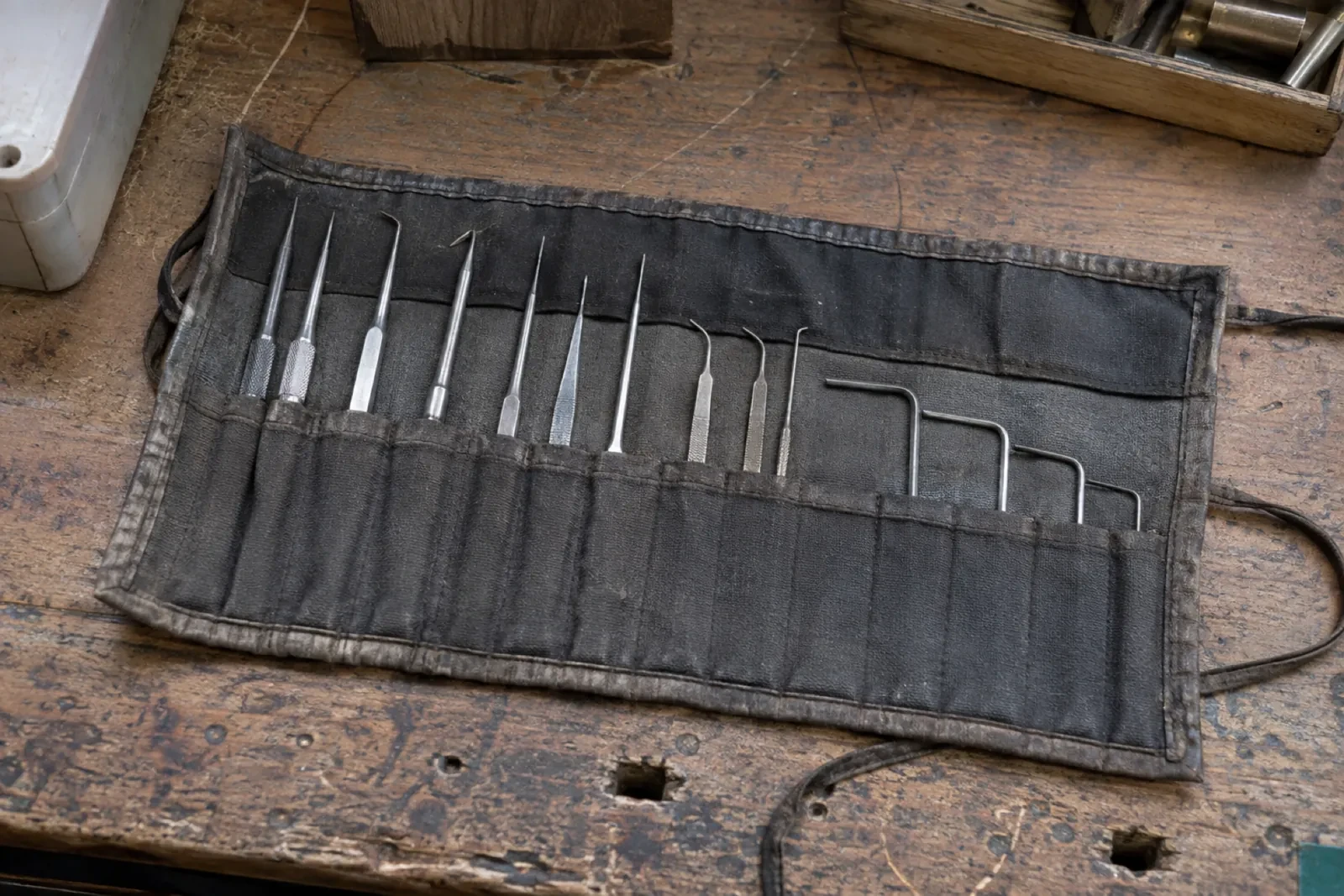 Roll-up black cloth pouch lying open on a worn wooden bench, holding thin metal probes and hooked tools in separate pockets.